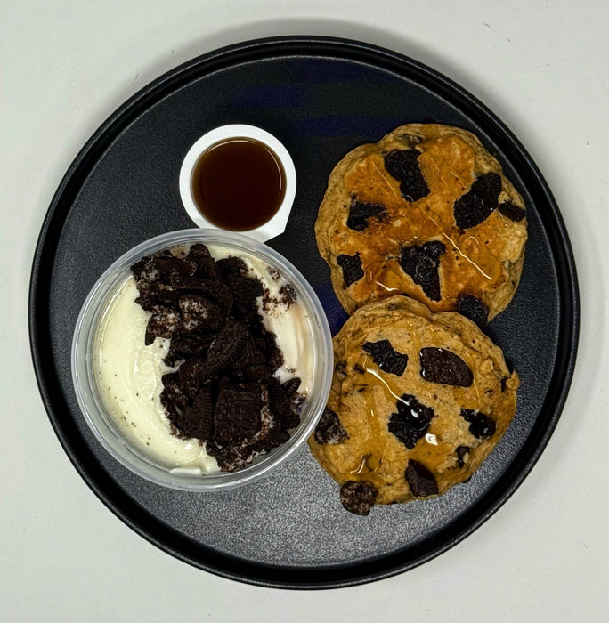 Two chocolate chip cookies on a black plate with a side of ice cream and syrup.