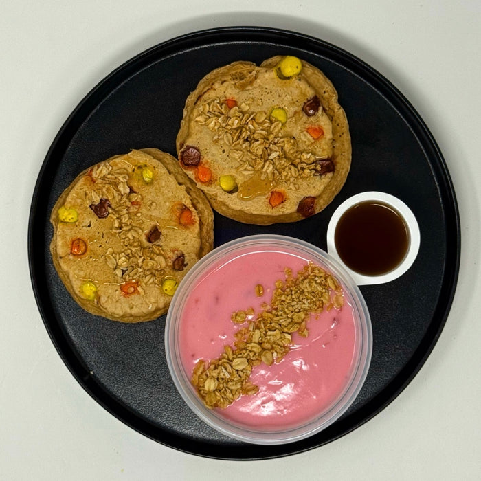 Two cookies with colorful toppings on a black plate, next to a container of pink yogurt with granola and a small bowl of syrup.
