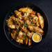 Overhead shot of loaded potato wedges in a black bowl, topped with crispy fried chicken, melted cheese, and a drizzle of yellow sauce, with a small side of mustard dipping sauce on a dark background.