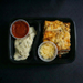Meal container with lasagna, mashed potatoes, and a side of sauce on a dark background