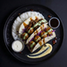 Overhead shot of a plated food dish on a black plate: two soft tacos filled with browned meat and green sauce, served with white rice, purple cabbage, two small dipping cups, and a yellow sauce smear on a dark background.