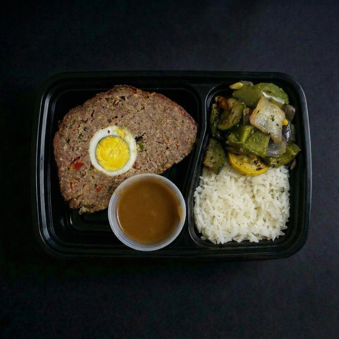 Meal in a black tray with meatloaf, egg, rice, and vegetables on a dark background