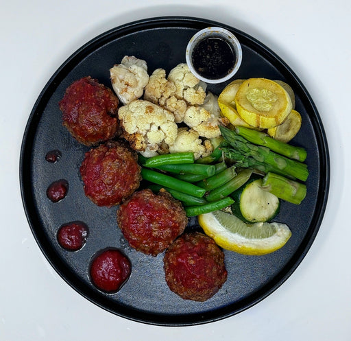 Black plate with meatballs, vegetables, and sauces on a white background.