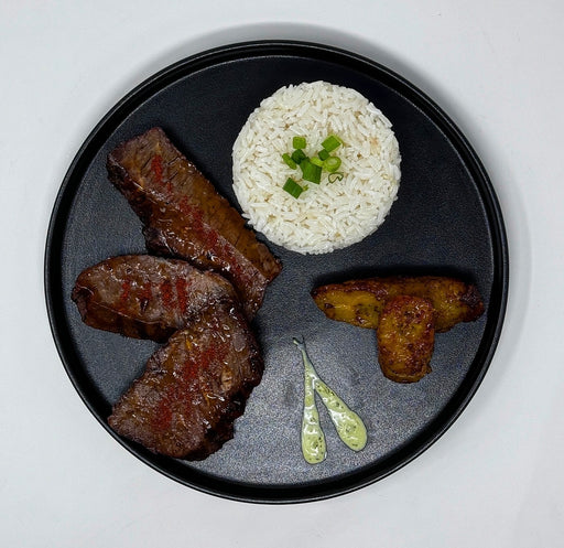 Black plate with grilled meat, white rice with green garnish, and fried plantains on a white background