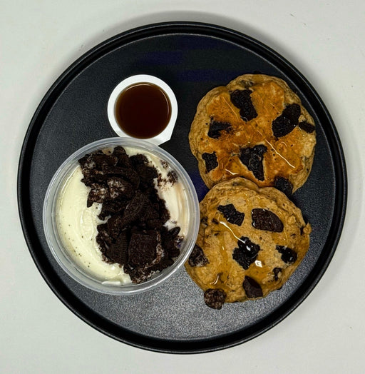 Two chocolate chip cookies on a black plate with a side of ice cream and syrup.