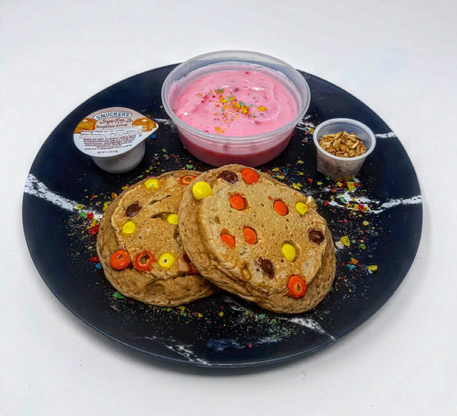 Stack of pancakes with colorful toppings on a black plate with side dishes including a pink yogurt and granola.