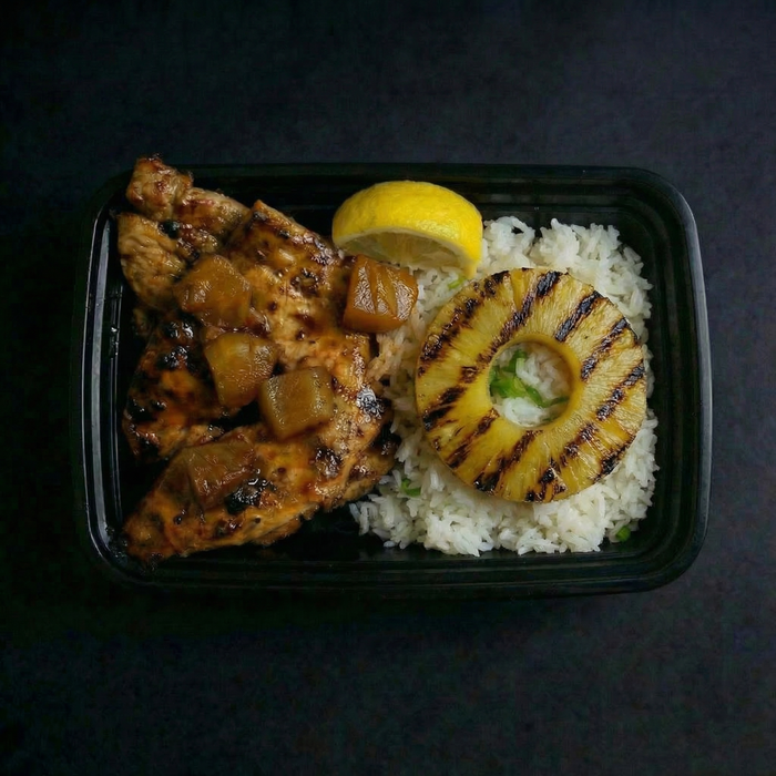 Grilled chicken with pineapple and rice in a black container on a dark surface.
