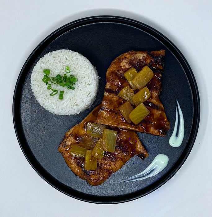 Grilled bbq chicken breast topped with pineapples and paired with jasmine rice and sauce on a black plate.