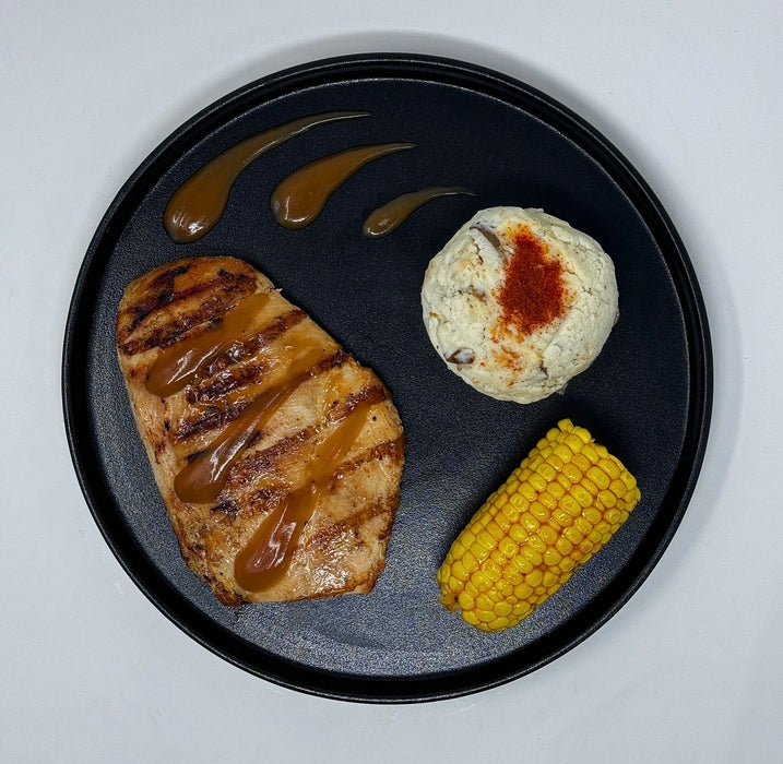 Plated dish with grilled meat, corn, and a round bread roll on a black plate.